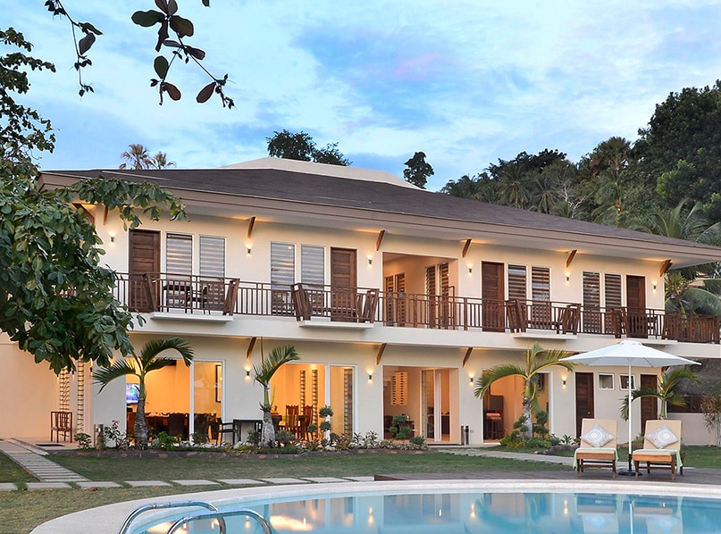 The villa's exterior is shown, featuring a two-story structure with multiple balconies. Lounge chairs sit beside a swimming pool, surrounded by well-maintained greenery. The setting is enhanced by a mix of natural and artificial lighting, reflecting a calm ambiance at dusk.
