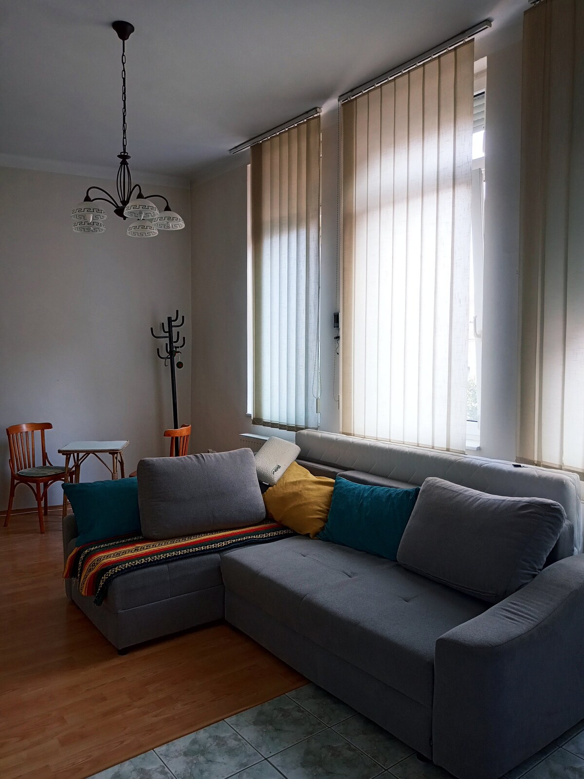 A light-filled living area is furnished with a grey sectional sofa adorned with colorful cushions. Two wooden chairs and a small table are positioned nearby. Vertical blinds filter natural light through the large windows, enhancing the spacious feel of the room.