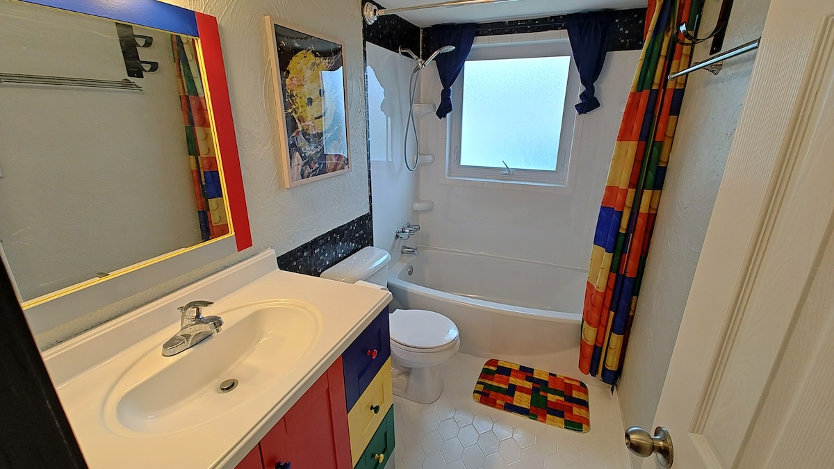 A colorful bathroom is shown, featuring a bathtub with a showerhead above. A white sink sits on a countertop with drawers painted in bright colors. A window provides natural light, while a patterned bath mat adds a playful touch to the floor.