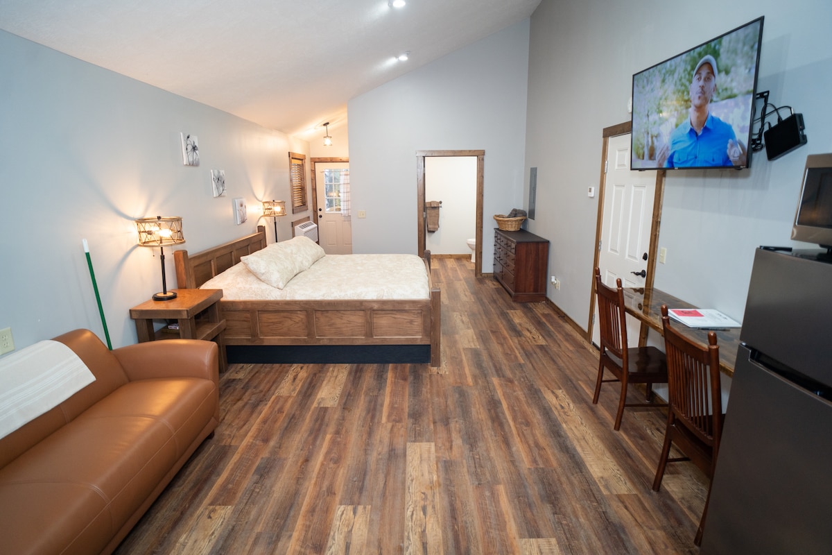 An open villa space features a spacious bed positioned adjacent to a leather couch. A large flat-screen TV is mounted on the wall, opposite a small dining area with two chairs and a table. The kitchenette and entry door are visible in the background.