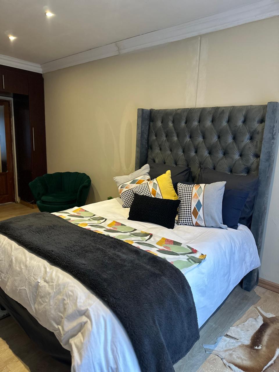 A spacious bed with a plush gray headboard is featured, adorned with colorful pillows and a decorative throw. A green accent chair is positioned nearby, while warm light illuminates the room, highlighting the soft textures and inviting atmosphere.