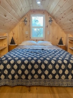 A cozy lofted bedroom features natural wood paneling and a large window allowing sunlight to fill the space. The spacious bed is dressed with a patterned blue and white blanket, complemented by simple wooden shelves on either side adorned with small decorative trees.