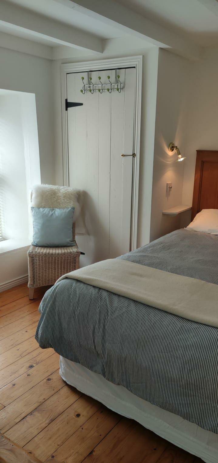 A cozy sleeping area features a spacious bed draped with a light blanket. Adjacent, a wicker chair is adorned with a soft, fluffy cushion. A small wardrobe stands in the corner with a single coat hook, and natural light filters through the window, illuminating the wooden floor.