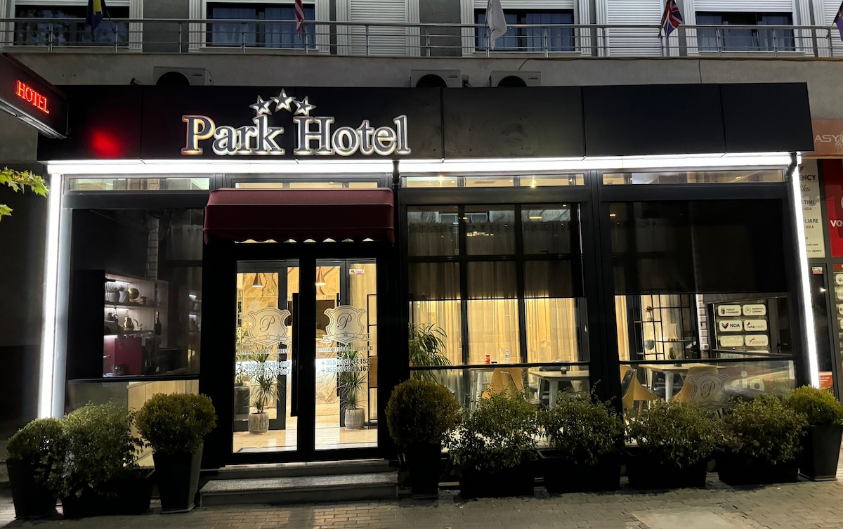 The hotel exterior features a modern facade with a prominent sign displaying 'Park Hotel.' Large windows offer views of the interior, which is illuminated warmly. Planters filled with greenery frame the entrance, enhancing the welcoming appearance.