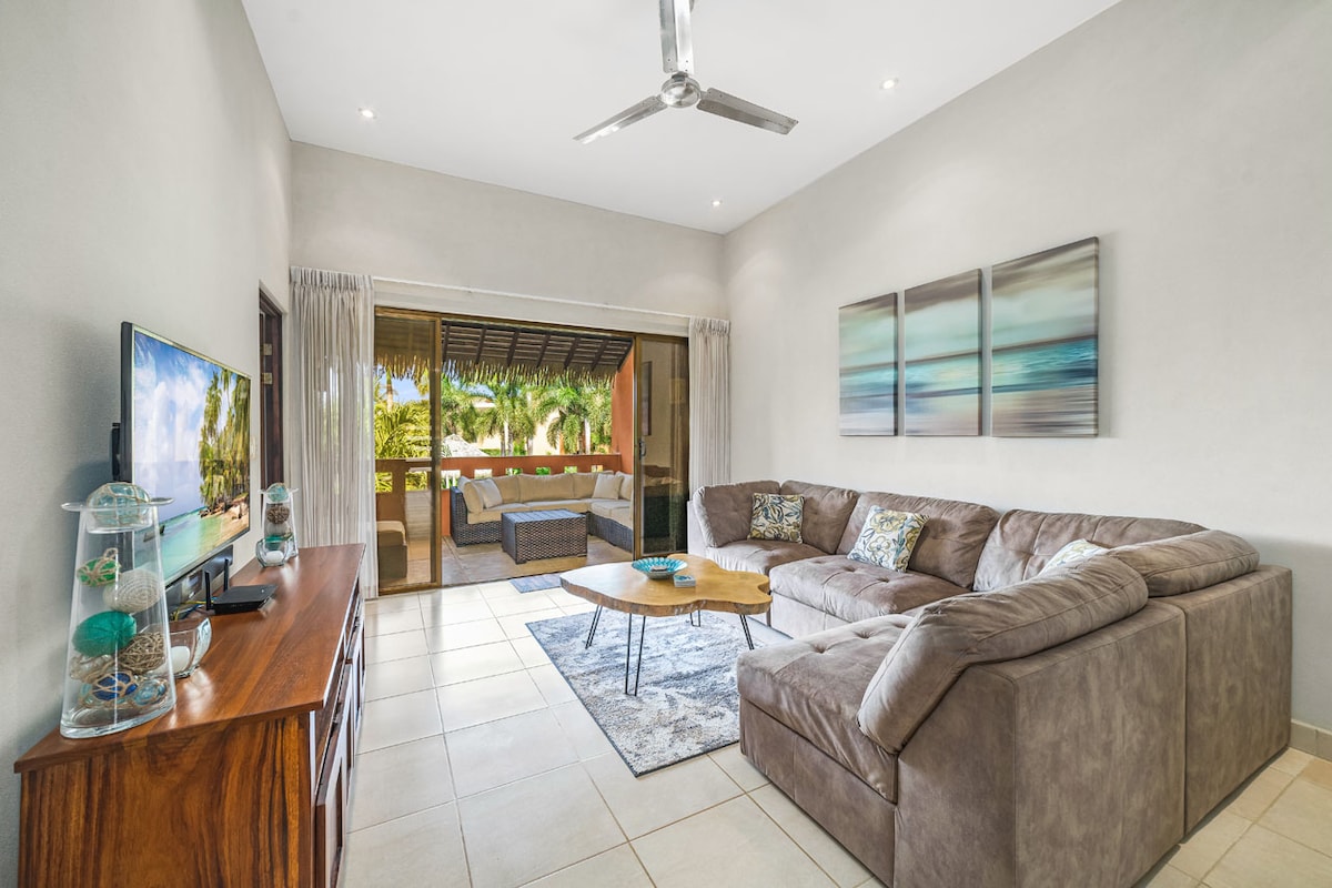 A spacious living area features a large sectional sofa arranged around a coffee table, creating a comfortable setting. A television is placed on a wooden media console. Large glass doors open to a balcony area, allowing natural light and views of the outdoors.