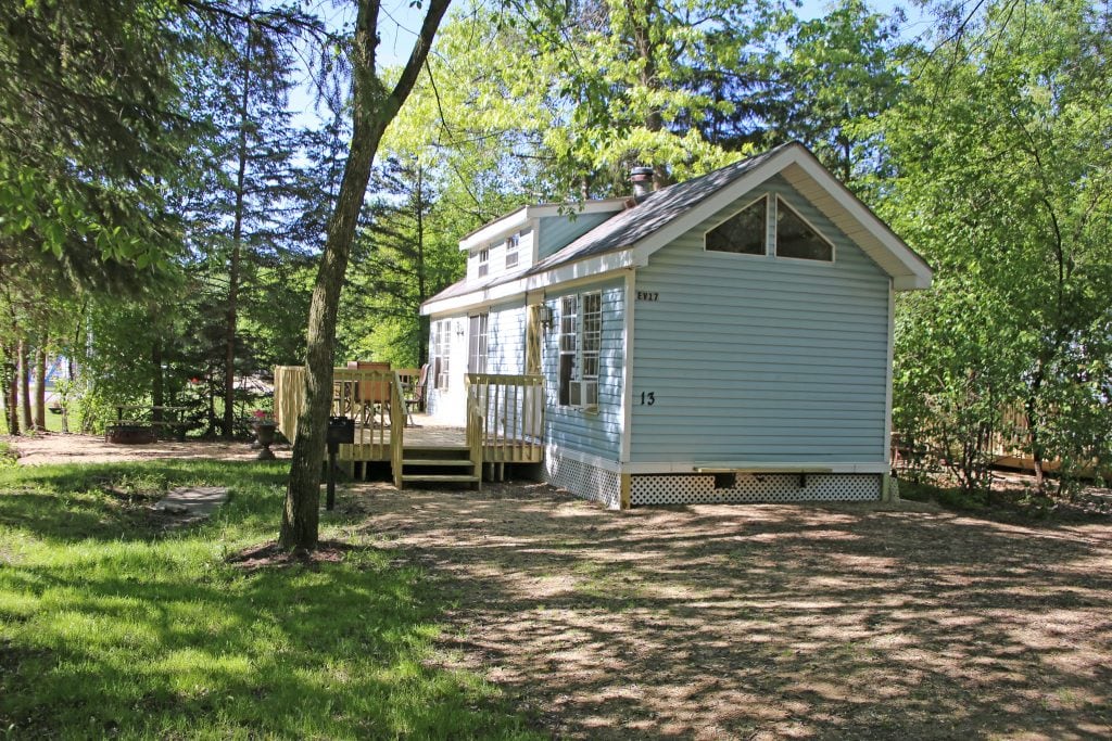 A light blue cabin is nestled among the greenery, surrounded by trees that provide a natural setting. A small wooden deck is visible at the front, offering outdoor access. Large windows allow for ample daylight, creating a bright and welcoming atmosphere.