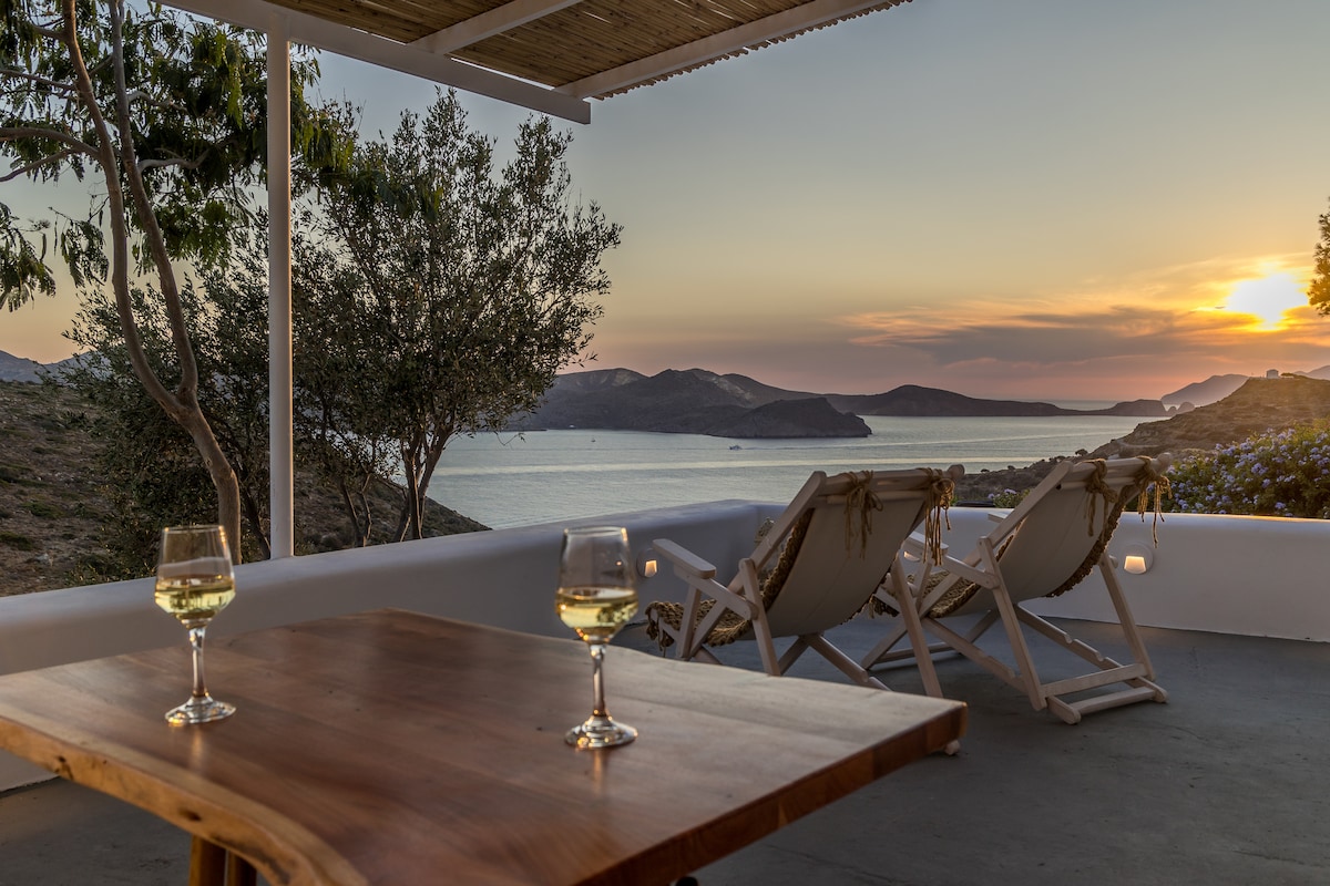 A spacious outdoor patio features two sun loungers and a wooden table set with two glasses. The backdrop includes a serene bay with distant hills, illuminated by a warm sunset sky blending soft oranges and purples.