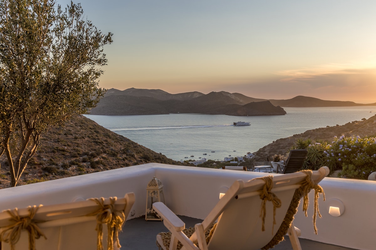 Two chairs are positioned on a patio, providing a serene view of Adamas Bay and the surrounding hills. The sunset casts a warm glow over the landscape, while a boat can be seen gently moving across the water.