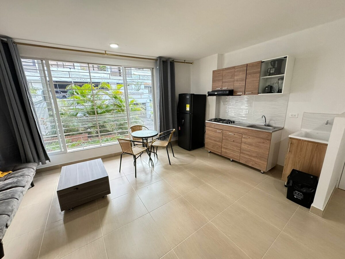 An open space is displayed, featuring a kitchen with wooden cabinetry and modern appliances. A dining area with a round table and two chairs is in the center. Large windows offer natural light, framed by grey curtains, and a small sofa is positioned nearby.