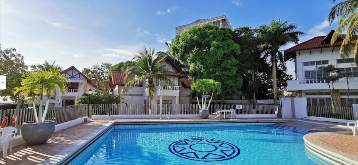 A clear blue swimming pool is surrounded by lush greenery and palm trees, with the sun reflecting on the water. Lounge chairs are positioned by the poolside. The backdrop features a multi-level residential building with balconies and large windows, adding to the spacious atmosphere.