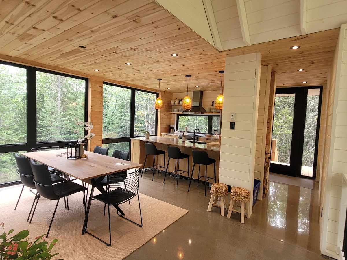 A modern dining area is featured with a wooden table surrounded by several black chairs. Large windows provide views of the surrounding forest. The kitchen area, positioned in the background, showcases bar stools and pendant lighting, enhancing the open-concept design.