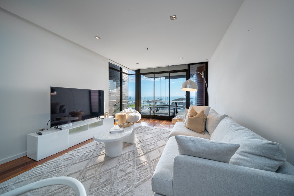A spacious living area features large glass doors, providing ample natural light and a view of the ocean. A light-colored sofa is paired with a round coffee table on a textured rug. The room offers a modern television mounted against the wall, creating a relaxed atmosphere.
