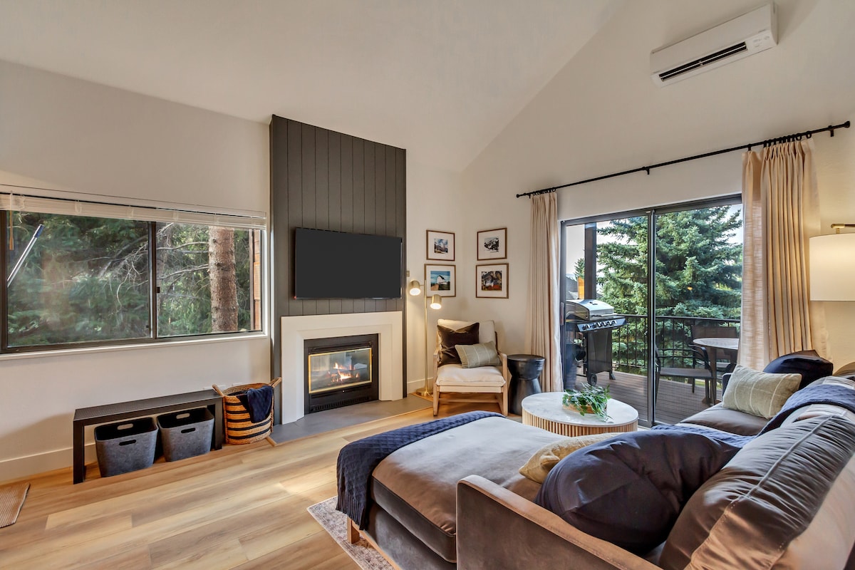 A cozy living area is highlighted by a gas fireplace set against a charcoal feature wall. Large windows invite natural light and showcase the outdoor trees. A plush sectional sofa is paired with a round coffee table and decorative plant, creating a comfortable seating space.