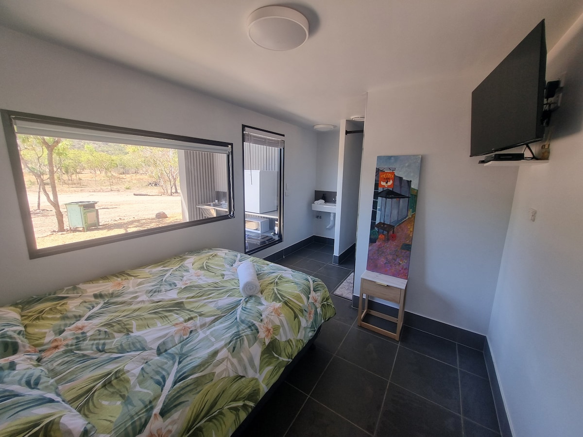 A cozy sleeping area is featured with a spacious bed adorned with a tropical patterned blanket. Natural light is allowed in through large windows. A mounted television and a colorful artwork provide visual interest, while the floor is covered with dark tiles.