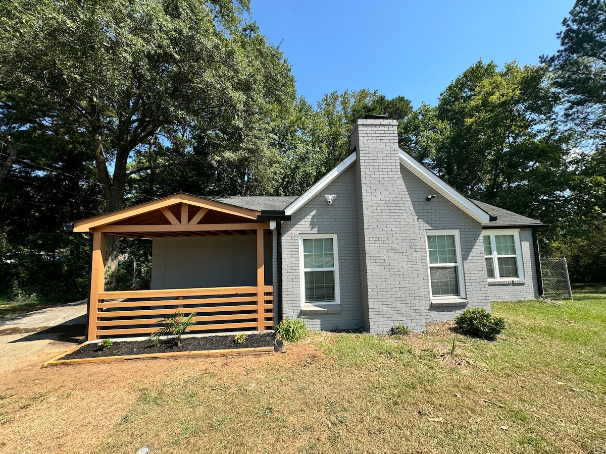 A single-story house exterior is shown, featuring a light gray brick facade and a welcoming porch with wooden accents. Lush greenery surrounds the property, while large windows provide a glimpse of the bright interior. A well-maintained lawn enhances the overall appeal.