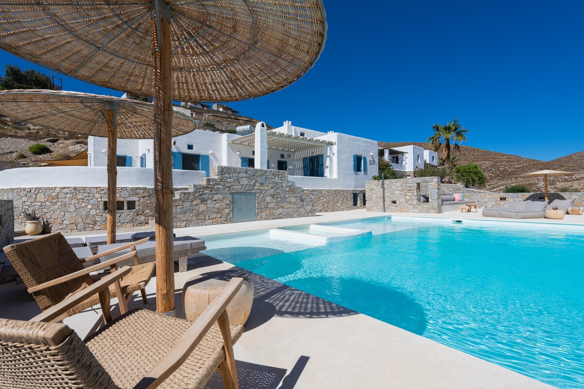 A private swimming pool is surrounded by elegant loungers and shaded by large parasols. The villa's white facade and blue shutters are visible in the background, set against a clear blue sky. Natural stone accents complement the tranquil pool area.