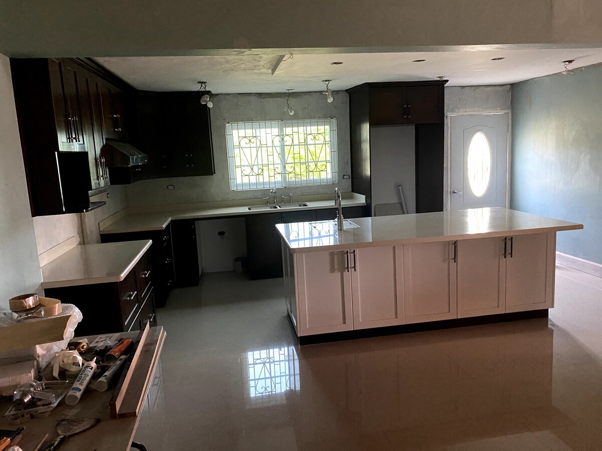 A spacious kitchen features dark wood cabinetry and a large white island in the center. Natural light filters through a window, illuminating the countertop. A door leads to the exterior, while the floor showcases a glossy finish.