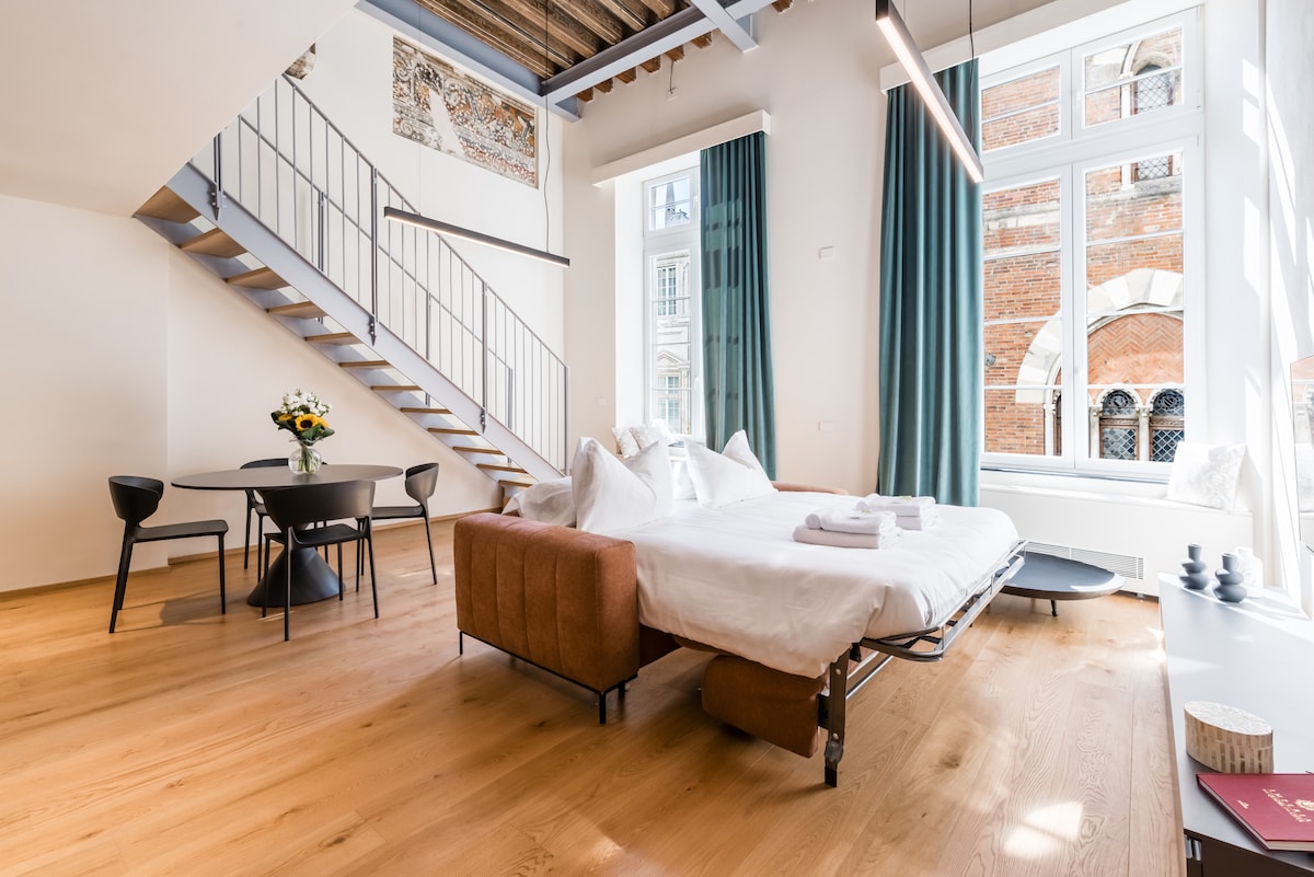 An open-plan living area features a spacious bed with fresh linens and pillows, along with a dining table for four. A stairway adorned with decorative frescoes leads to the lofted sleeping area. Large windows provide natural light and a view of the street below.
