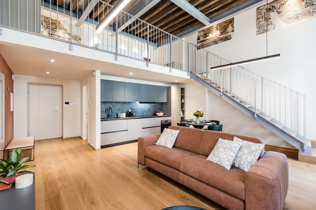 A spacious living area features a comfortable sofa adorned with decorative cushions. The open layout showcases a modern kitchen with sleek cabinetry, and wooden beams are visible overhead. A staircase leads to the lofted sleeping area, adding a unique architectural element to the room.