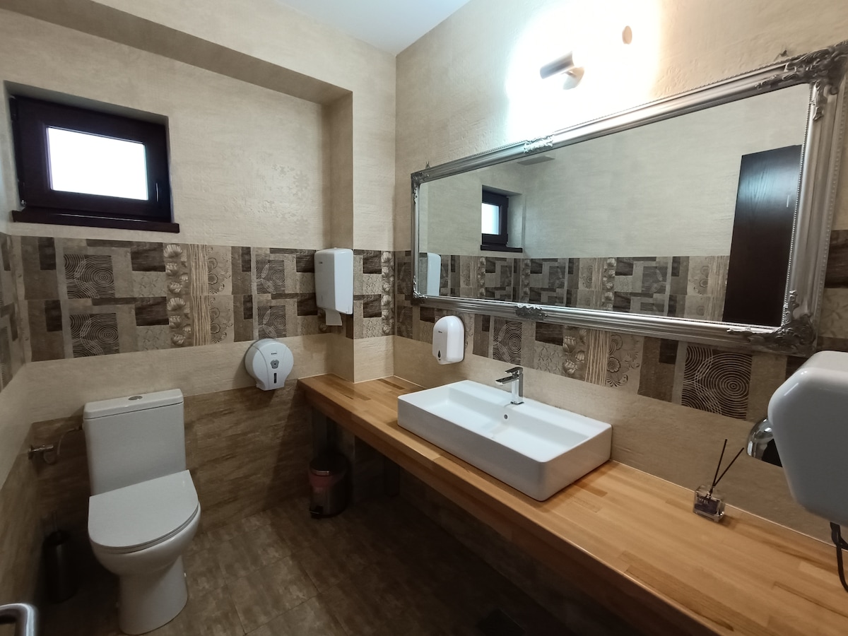 A modern bathroom features a spacious wooden countertop with a rectangular sink and a large mirror above. The walls are adorned with a mix of textured tiles, while minimalistic soap dispensers and hand dryers are mounted for convenience. Natural light enters through a small window.