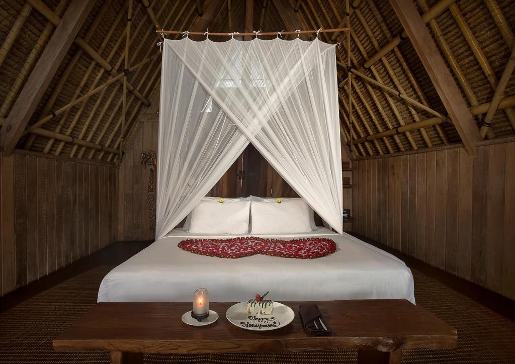 A spacious bedroom features a four-poster bed adorned with white drapes and a decorative arrangement of petals. A wooden table is positioned nearby with a candle and a small cake. The room showcases natural wooden walls and a thatched ceiling.