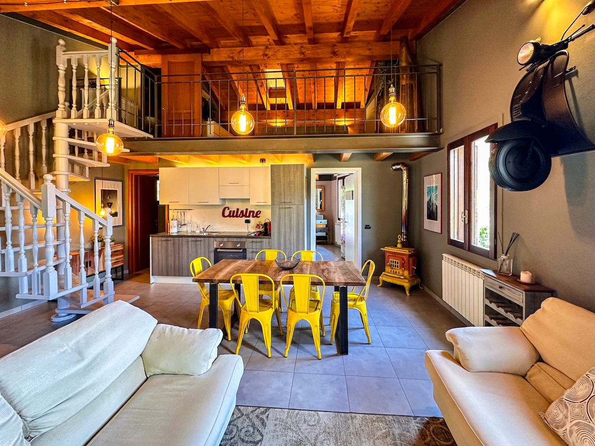 The open living space features a wooden loft above, accentuating the high ceilings. A large dining table surrounded by yellow chairs is positioned centrally. The kitchen, equipped with modern appliances, is visible in the background, complemented by natural light from multiple windows.