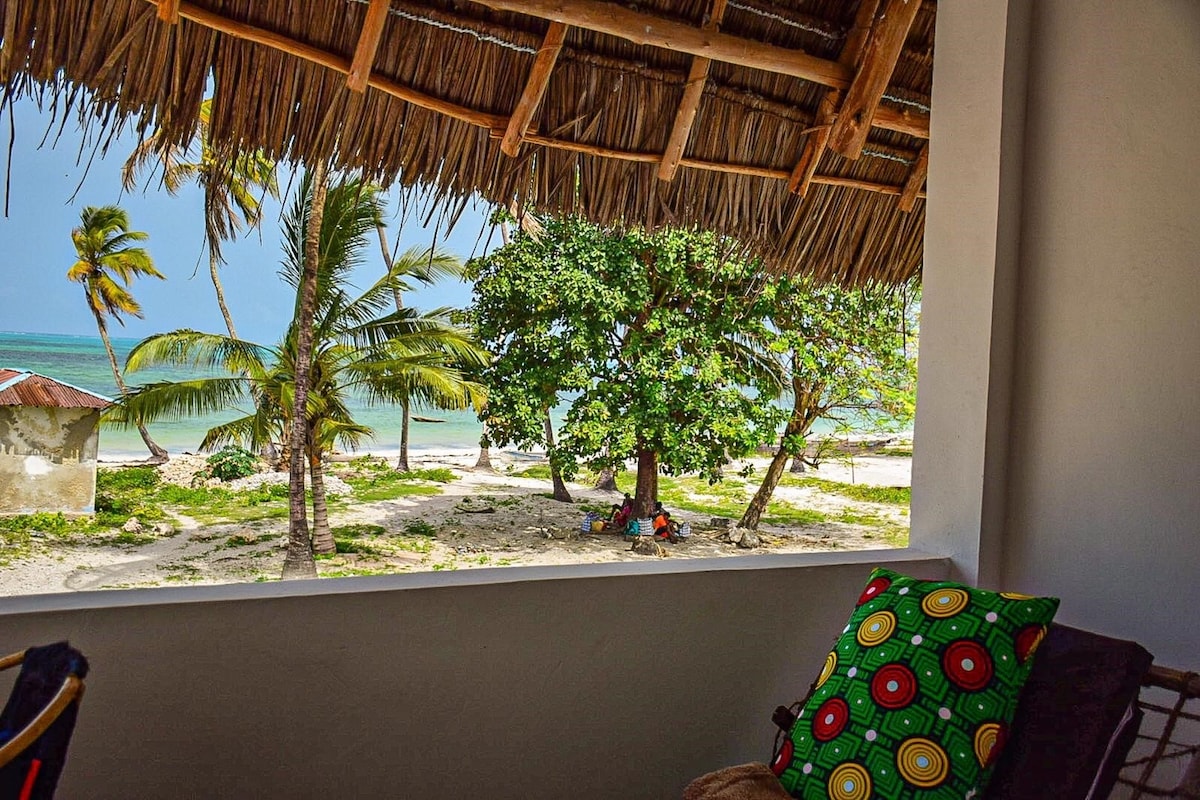 A view from the terrace showcases swaying palm trees and a sandy beach leading to turquoise waters. A thatched roof provides shade to the seating area, while a colorful cushion adds a touch of comfort to the setting.