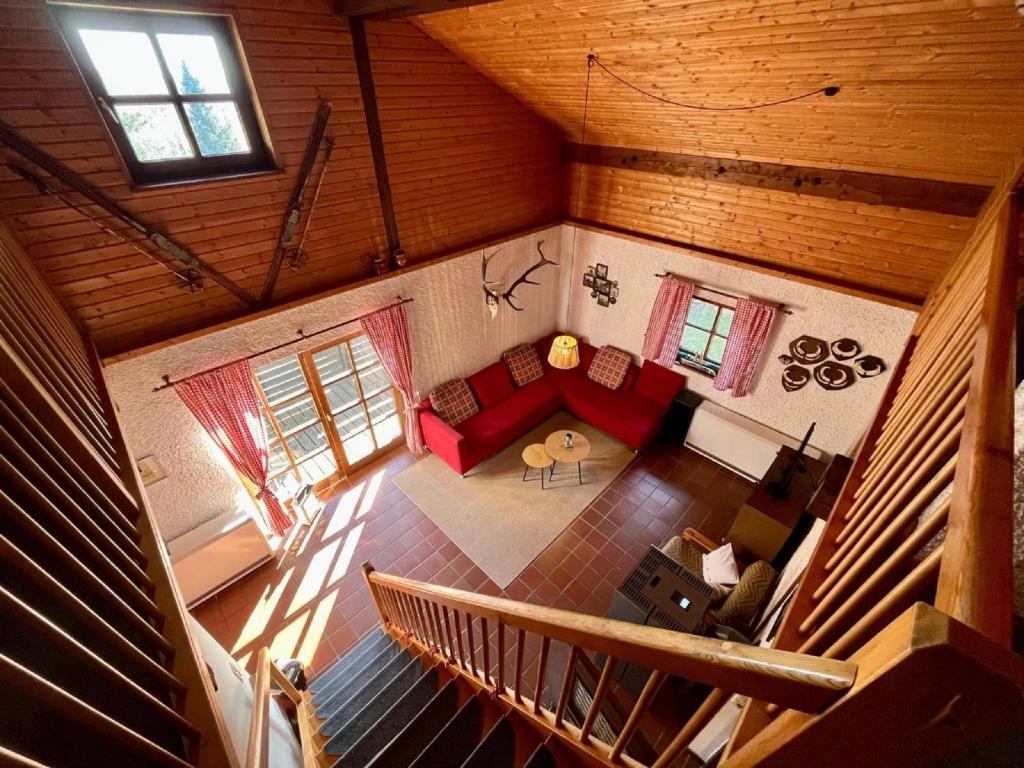 The spacious living area features wooden beams and a warm color palette. A red sectional sofa is positioned around a circular coffee table, while large windows allow natural light to fill the room. A staircase with wooden railings leads to the upper level.