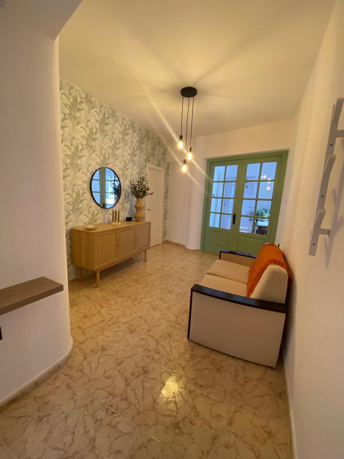 A hallway features light-colored flooring and a patterned wall with leafy designs. A stylish console table is positioned against one wall, accompanied by a round mirror. Natural light streams through a set of green-framed double doors leading outside, while a small seating area includes throw pillows.