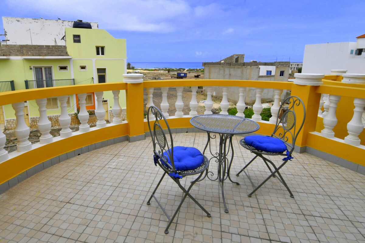 An outdoor terrace is featured, furnished with a small round table and two wrought iron chairs, each with blue cushions. The railing is adorned with decorative columns, offering a view of the surrounding landscape and distant sea.
