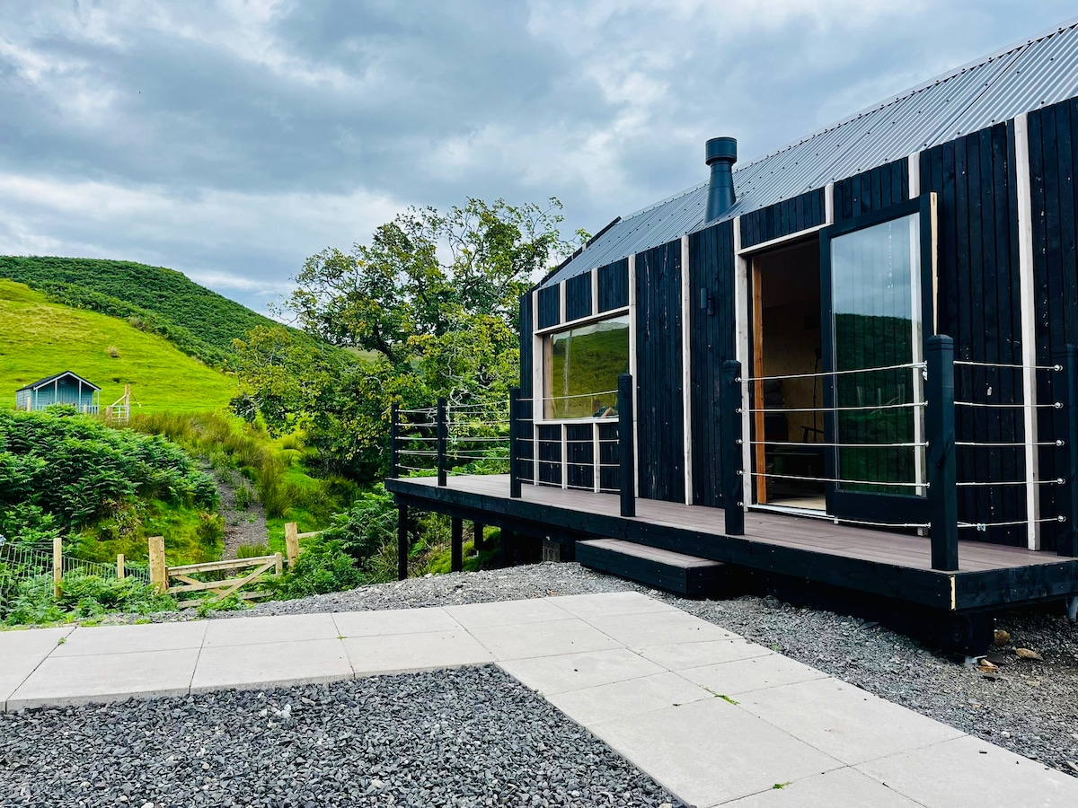 A modern wood-clad bothy is featured with large glass doors opening onto a spacious deck. Surrounding greenery and hills provide a natural backdrop. A gravel pathway leads to the entrance, emphasizing the secluded and tranquil setting of the property.
