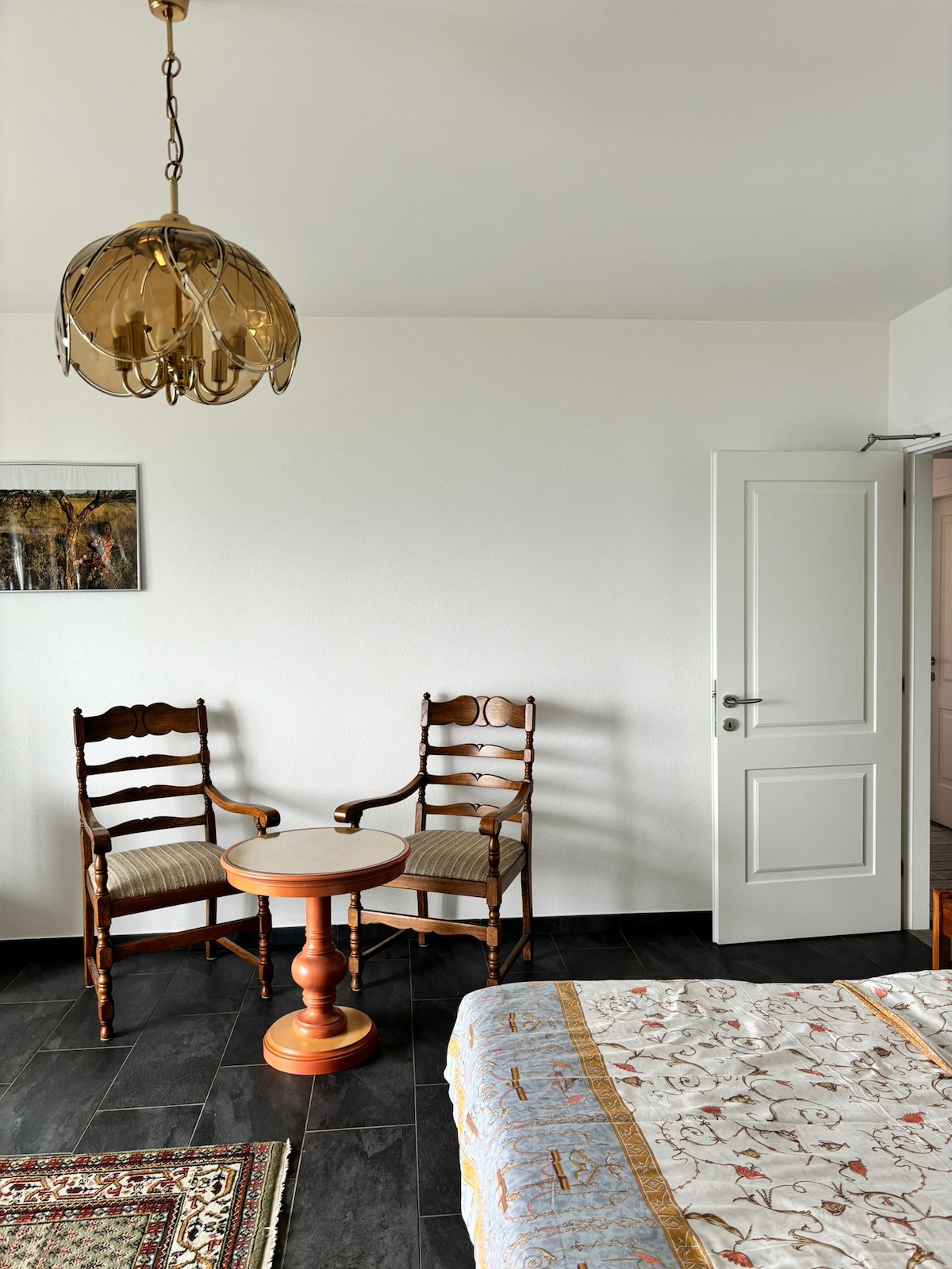 A cozy corner features a round table surrounded by two wooden chairs with upholstered seats. Above, a decorative light fixture hangs from the ceiling. The space is bright and accented by a small area rug on the dark tile floor.