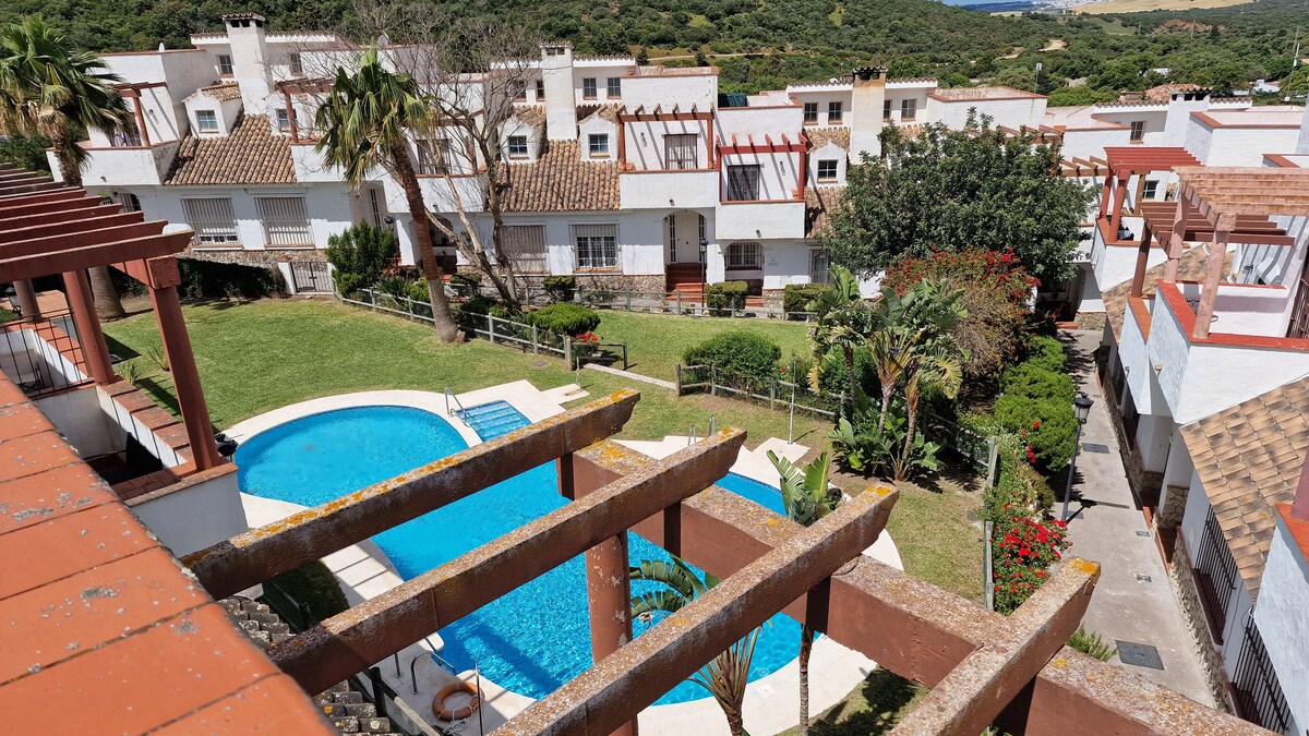 A spacious outdoor area is visible, featuring a blue swimming pool surrounded by lush greenery. Several buildings with tiled roofs and balconies are in the background, alongside palm trees and flowering plants, creating a serene environment.