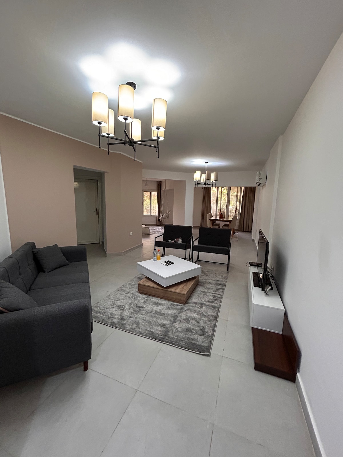 A modern living area is presented with a grey sofa and two black chairs surrounding a low coffee table, featuring a rug beneath. Natural light filters in from tall windows, while a chandelier and wall-mounted TV add contemporary accents.