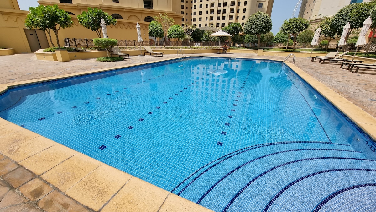 A tranquil outdoor pool occupies the center of the image, surrounded by lush greenery and well-maintained lounge chairs. Sun umbrellas are positioned nearby, offering shaded areas for relaxation. The clear blue water reflects the surrounding environment under bright daylight.