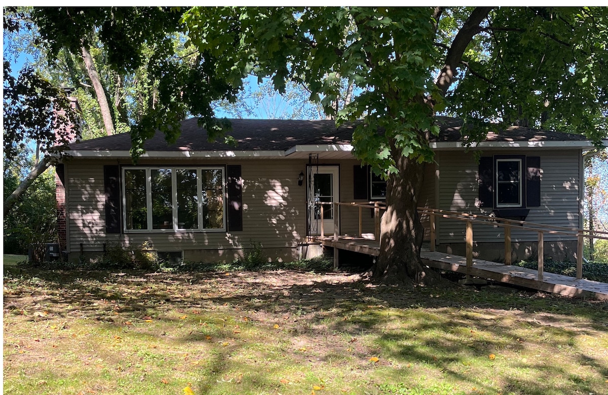 A single-story home is situated among lush trees, featuring a gently sloping front yard. A wooden ramp leads to the entrance, ensuring accessibility. Large windows allow natural light to fill the living space, while the backdrop of vibrant greenery adds to the serene surroundings.