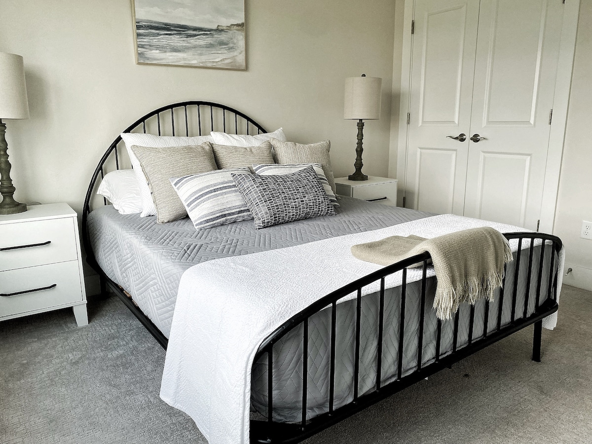 A comfortable bedroom features a spacious bed adorned with various throw pillows and a light blanket. Two bedside tables with lamps flank the bed, and a piece of nature-inspired artwork is hung above. Soft carpet underfoot complements the neutral wall colors.
