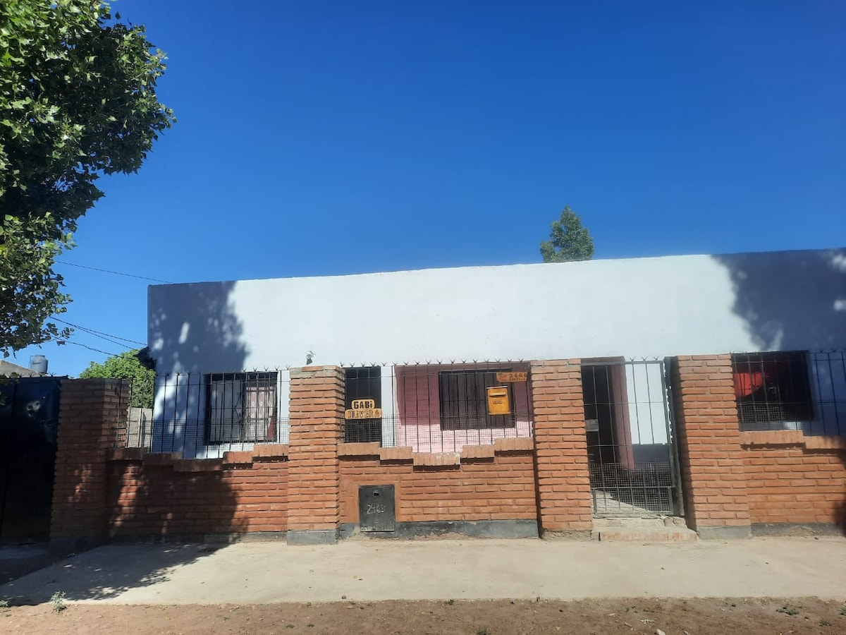 A single-story building is shown with a brick exterior and a white facade. Metal bars are present on the windows, and a clear blue sky serves as the backdrop. A dirt pathway leads to the entrance, surrounded by sparse vegetation.