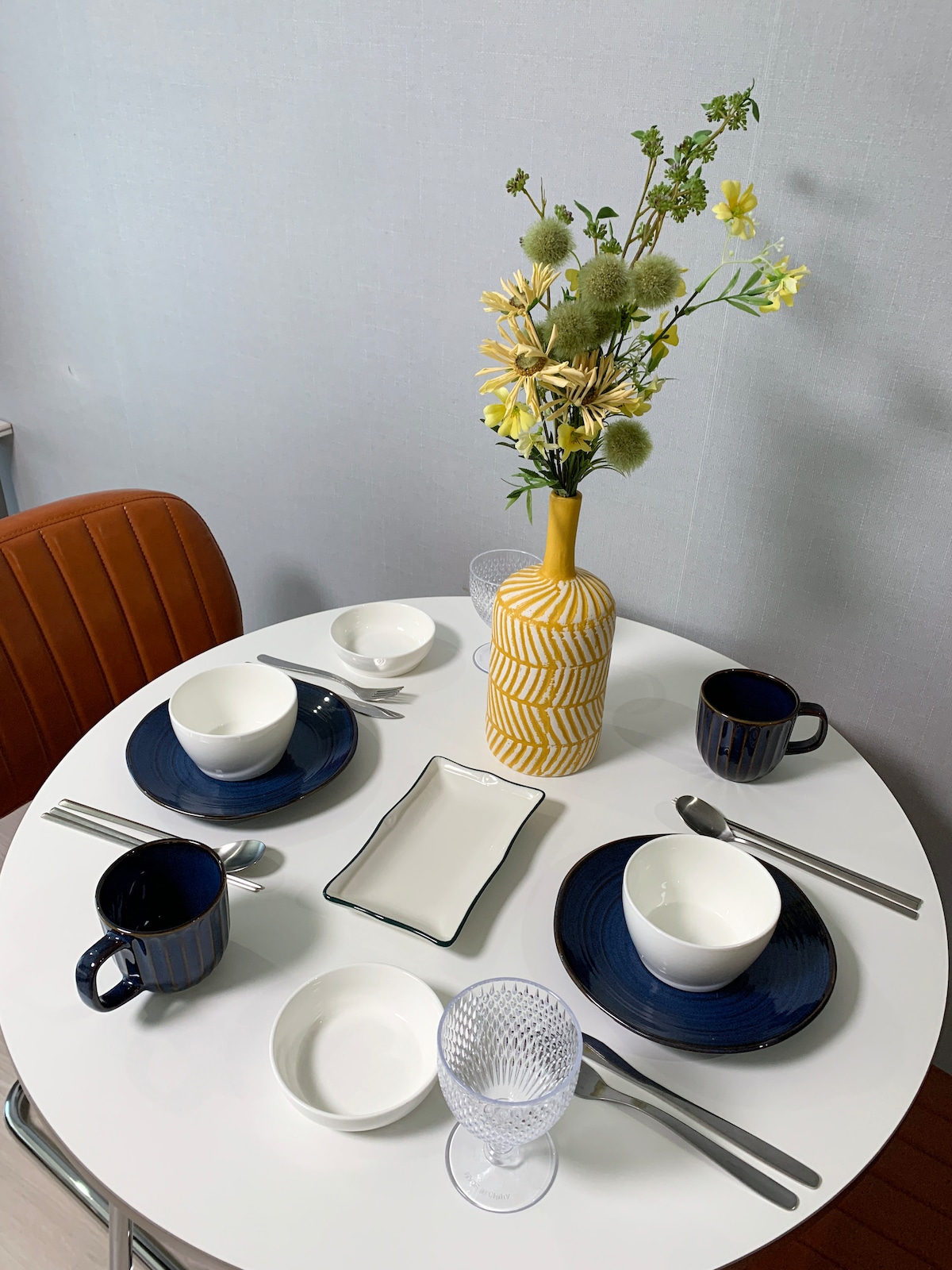 A dining table set for two showcases a minimalist arrangement. Two plates, bowls, and cups are placed neatly alongside utensils. A decorative vase filled with flowers serves as a centerpiece, complemented by a rectangular serving dish and a glass.