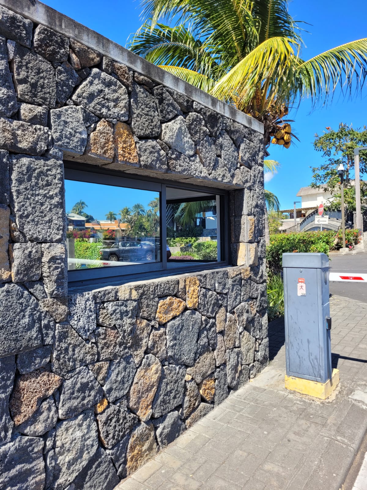 A rustic stone structure features a large window framed in black, allowing a view of the surroundings. Tropical palm trees are visible in the background, contributing to the coastal atmosphere. The well-maintained pathway leads to the entrance of the complex.