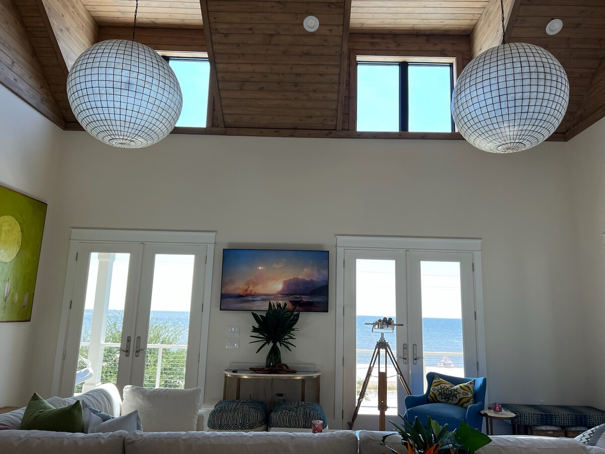 The living area features two large pendant lights hanging from a vaulted ceiling, with ample natural light streaming in through two wide French doors. A scenic view of the beach is framed by the doors, and a colorful artwork accentuates the wall.