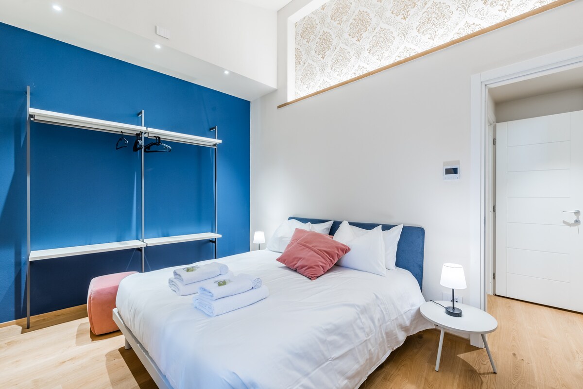 A well-appointed bedroom features a spacious bed with crisp white linens and a decorative pillow. A clothing rack is positioned against a vibrant blue wall, complemented by warm wooden flooring. Soft lighting from bedside lamps creates a relaxed atmosphere.