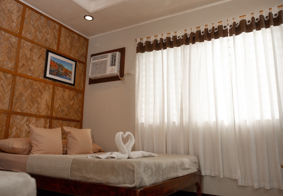 A cozy sleeping area features a spacious bed with beige linens and two decorative pillows. A white towel is folded into a swan shape on the bed. Natural light filters through a set of sheer white curtains, illuminating the bamboo wall and providing a warm ambiance.