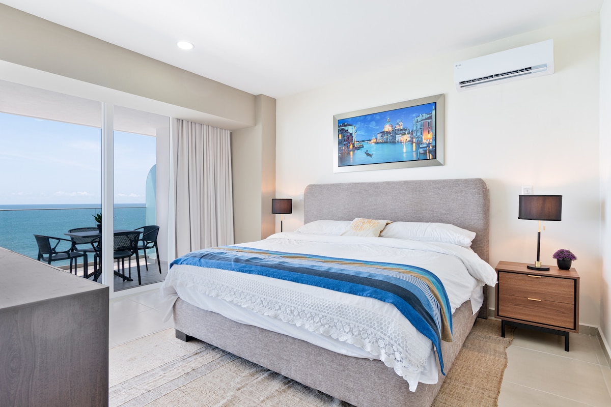 A spacious bedroom features a large bed dressed in white linens with a blue-accented blanket. Two lamps sit on bedside tables beside the bed. A view of the ocean is visible through expansive glass doors leading to a private balcony.