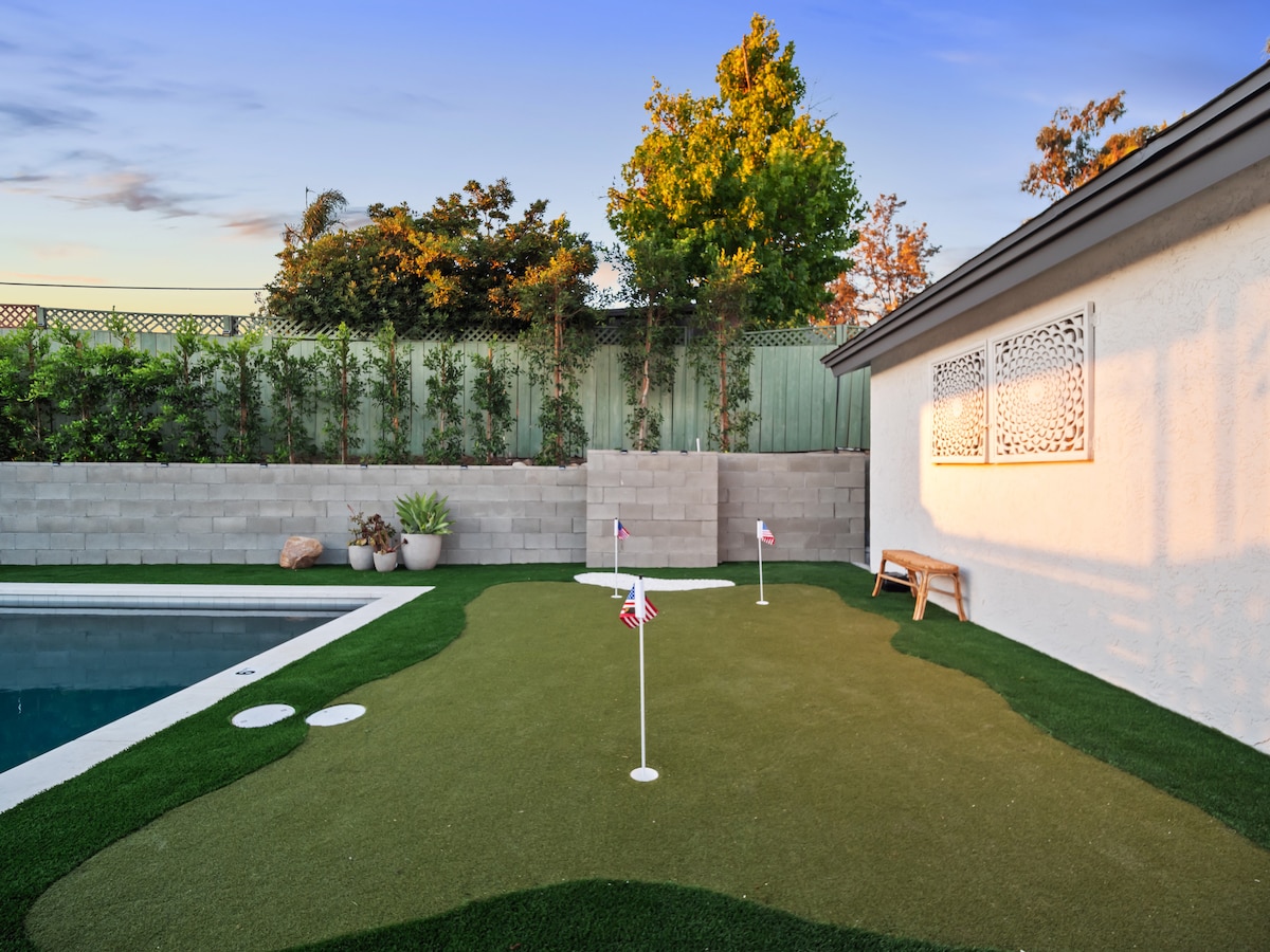 A well-maintained backyard features a golf putting green with multiple holes and flags planted in the turf. A refreshing pool is visible nearby, surrounded by lush greenery and a concrete wall, offering a serene outdoor space for relaxation and entertainment.