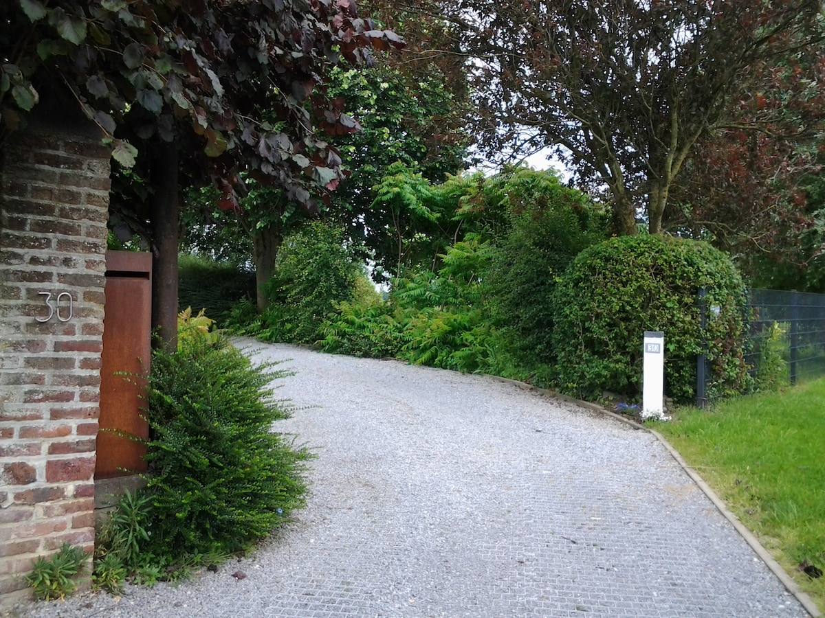 A gravel driveway leads through lush greenery, framed by well-maintained shrubs and trees. A brick column and a decorative post provide welcoming entry points, while the overall scene conveys a sense of peace and natural beauty.