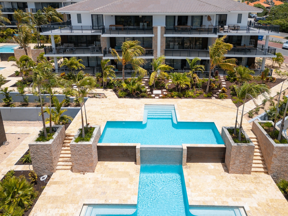A tranquil outdoor area features several pools surrounded by landscaped gardens. The space is designed with natural stone accents and palm trees, offering a serene environment. Multi-level balconies of the apartments overlook the pools, creating an inviting atmosphere for relaxation.