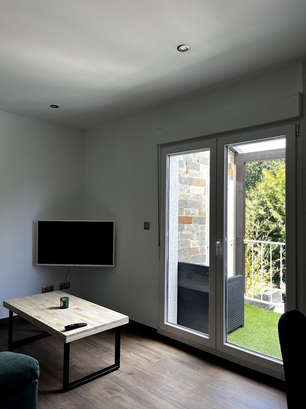 A living area features a modern flat-screen television mounted on the wall. A wooden coffee table sits in front of a plush sofa. Light enters through glass doors, which lead to a small balcony with greenery visible outside.