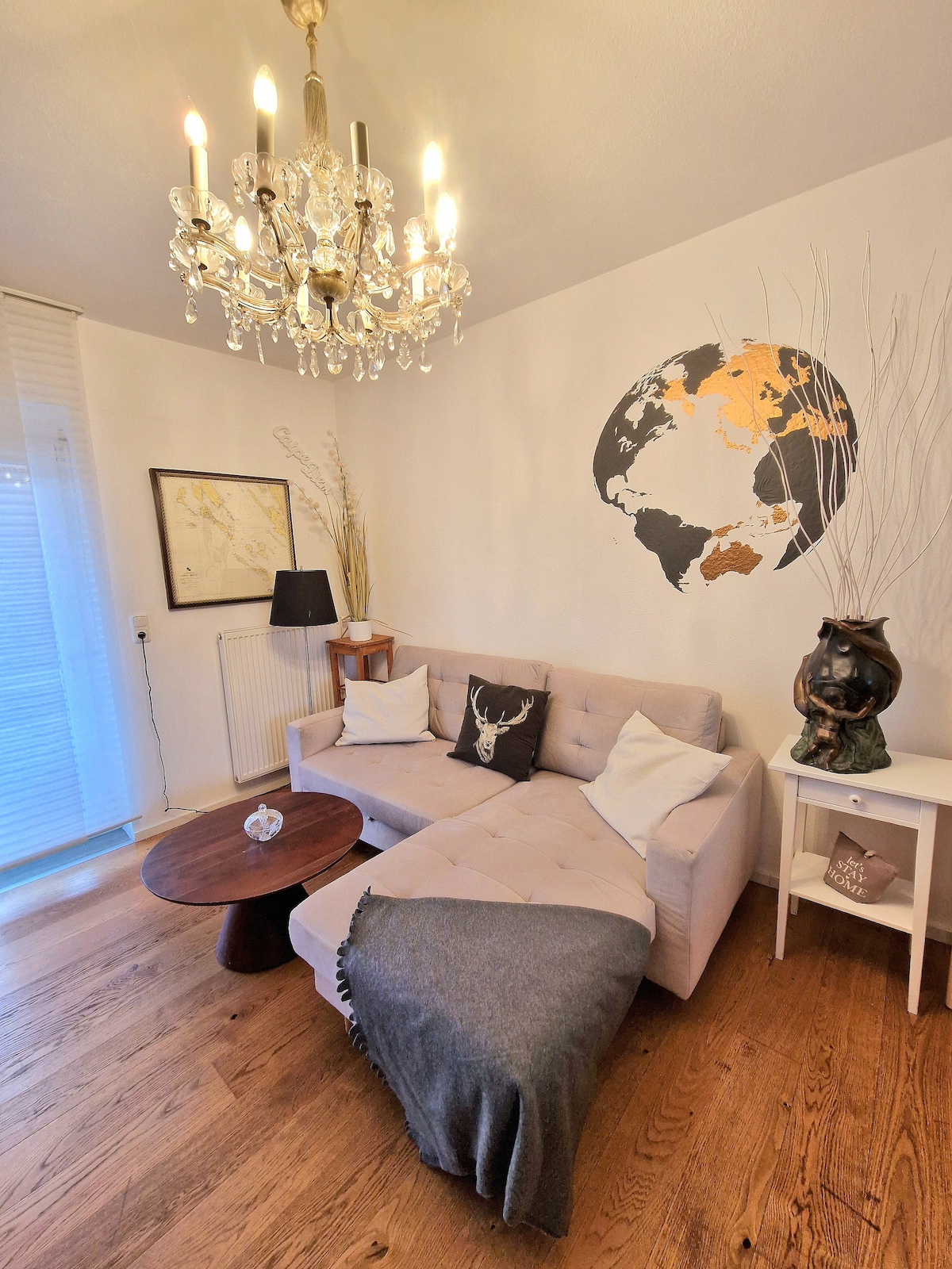 A cozy living area features a light-colored sectional sofa adorned with cushions. A small round table sits nearby, accompanied by a decorative lamp and an art piece depicting a world map. Natural light filters through sheer curtains, highlighting the wooden floor.