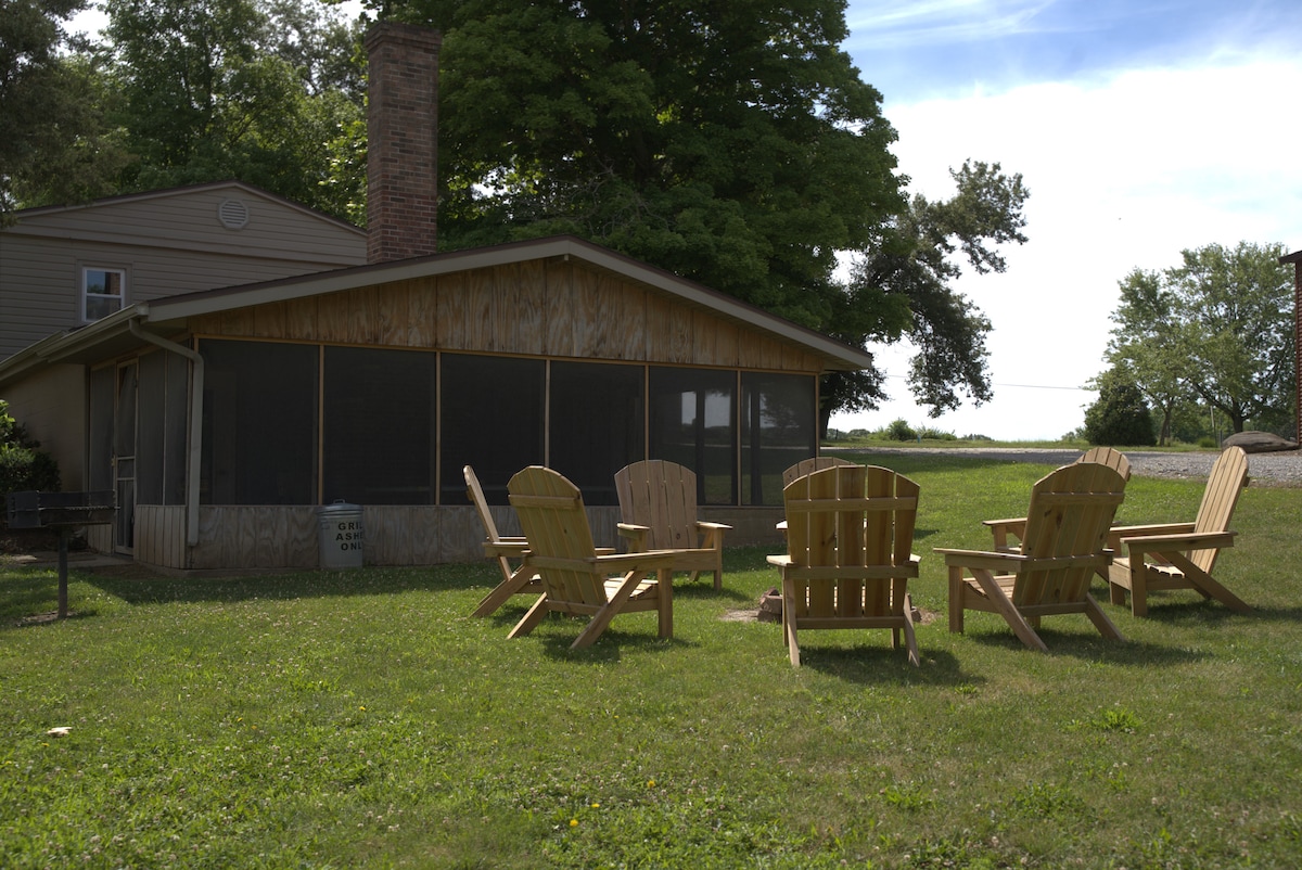 A spacious outdoor area features five wooden Adirondack chairs arranged around a fire pit, set on a well-maintained grassy lawn. A screened porch is visible in the background, attached to a house surrounded by mature trees, creating a peaceful retreat.